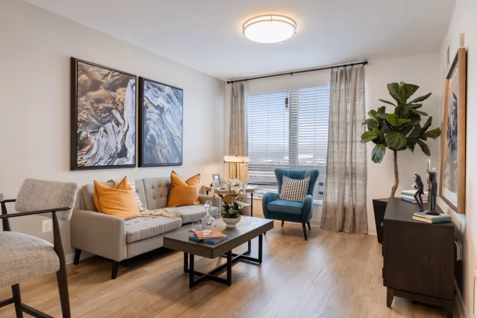 A modern living room with a light gray sofa adorned with orange and patterned pillows, a blue armchair with a black and white pillow, a wooden coffee table with books and decorative items, two abstract art pieces on the wall, a large window with blinds and sheer curtains, a tall green plant in a black pot, and a dark wooden sideboard with sculptures and books.