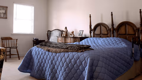 A bedroom with a wooden bed covered by a blue quilt, a dresser with figurines, chairs, and a window.