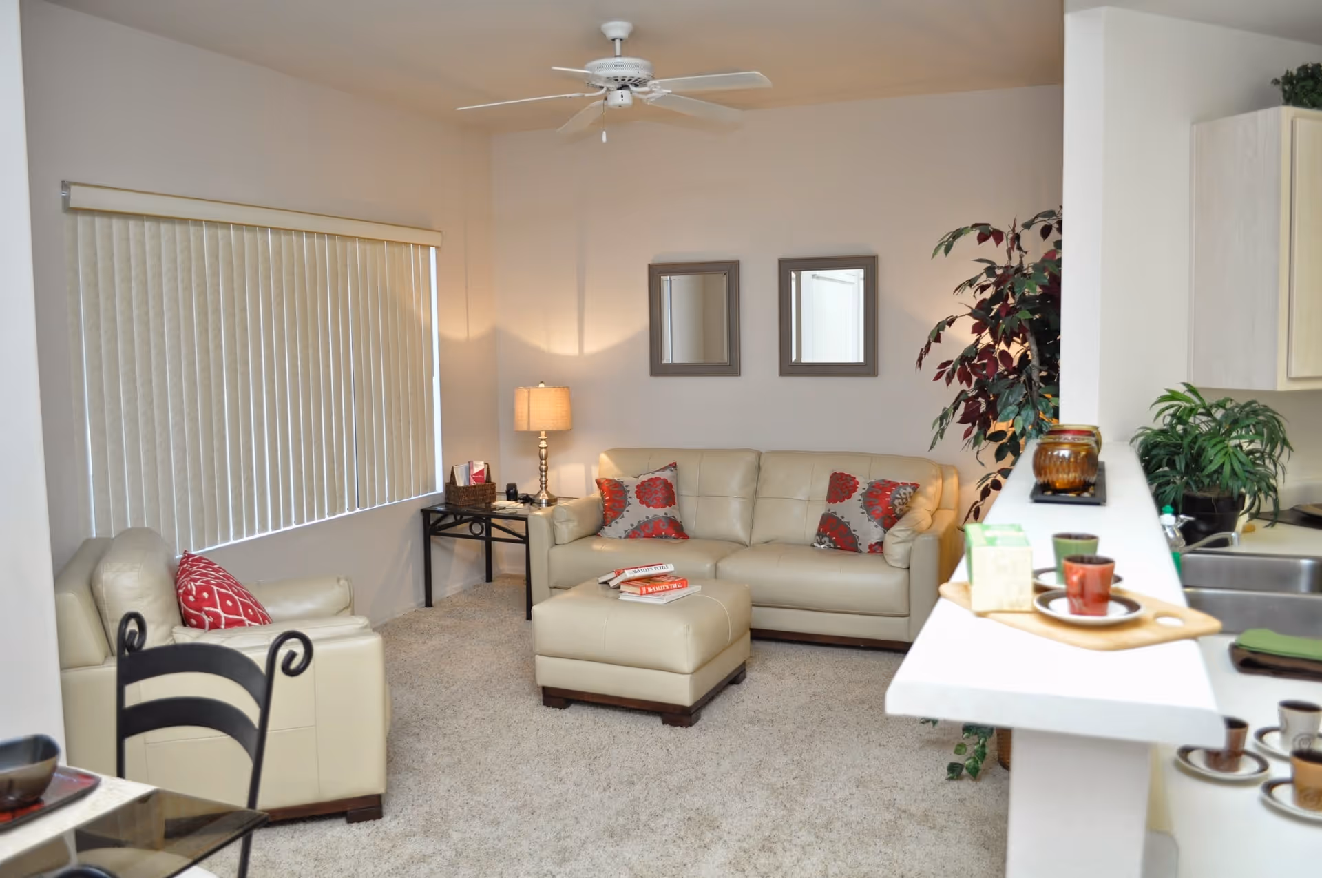 A cozy living room with beige leather sofa and armchair, decorated with red and gray patterned pillows. There is a matching ottoman in front of the sofa with books on it. The room has beige carpet, a ceiling fan, two framed mirrors on the wall, a side table with a lamp, and a large window with vertical blinds. A kitchen counter with cups and plants is visible on the right side.