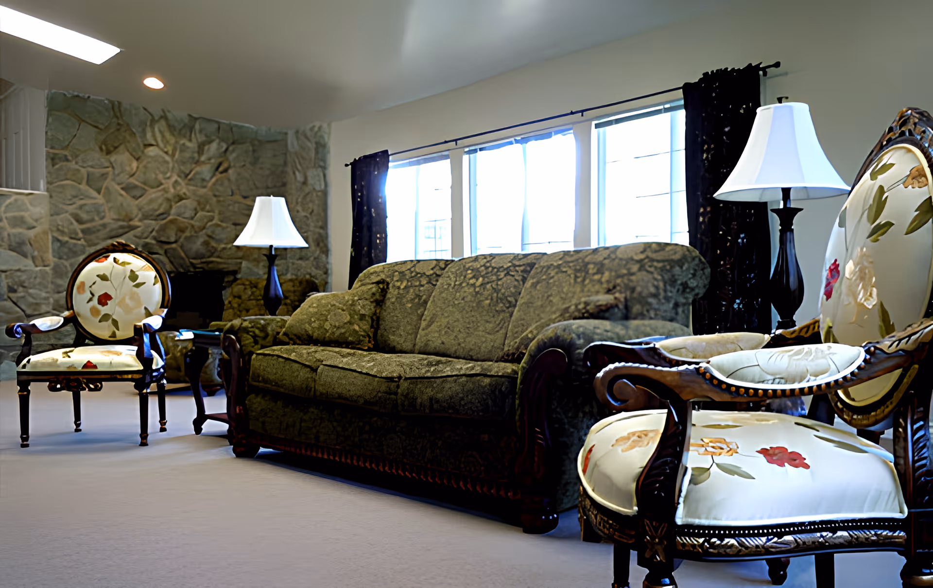 A living room with a green patterned sofa, two floral upholstered armchairs with wooden frames, two table lamps with white shades, large windows with dark curtains, and a stone wall with a fireplace.