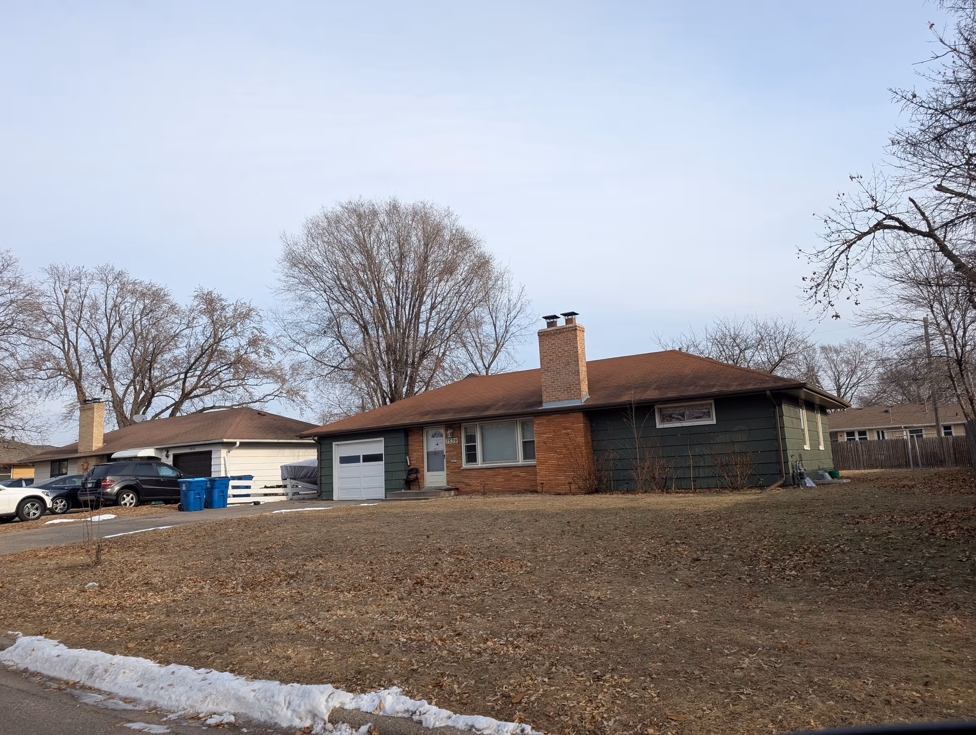 A single-story residential house with a brown roof, green siding, and a brick chimney. The house has a garage with a white door and a driveway with several parked cars. Leafless trees are visible in the background, and there is a patchy lawn with some snow along the street curb.