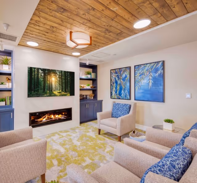 A cozy living room with beige armchairs featuring blue patterned cushions, a modern fireplace beneath a wall-mounted TV displaying a forest scene, built-in shelves with plants and books, two large blue and green nature-themed paintings on the wall, a light green and beige patterned rug, and a wooden ceiling with recessed lighting.