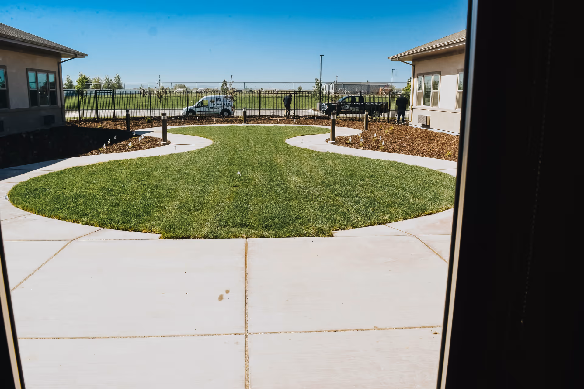 View through a window of a courtyard area with a green lawn and curved concrete pathways, bordered by two single-story buildings on either side. In the background, there is a black metal fence, two vehicles, and a few people near the fence under a clear blue sky.