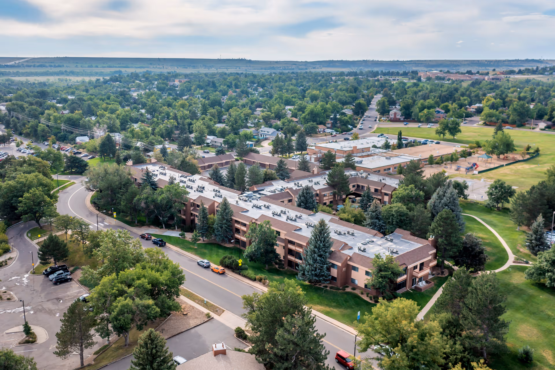 Aerial view of Brookdale Meridian Boulder senior living facility surrounded by trees and greenery, with nearby roads, parking areas, and a playground visible in the background.