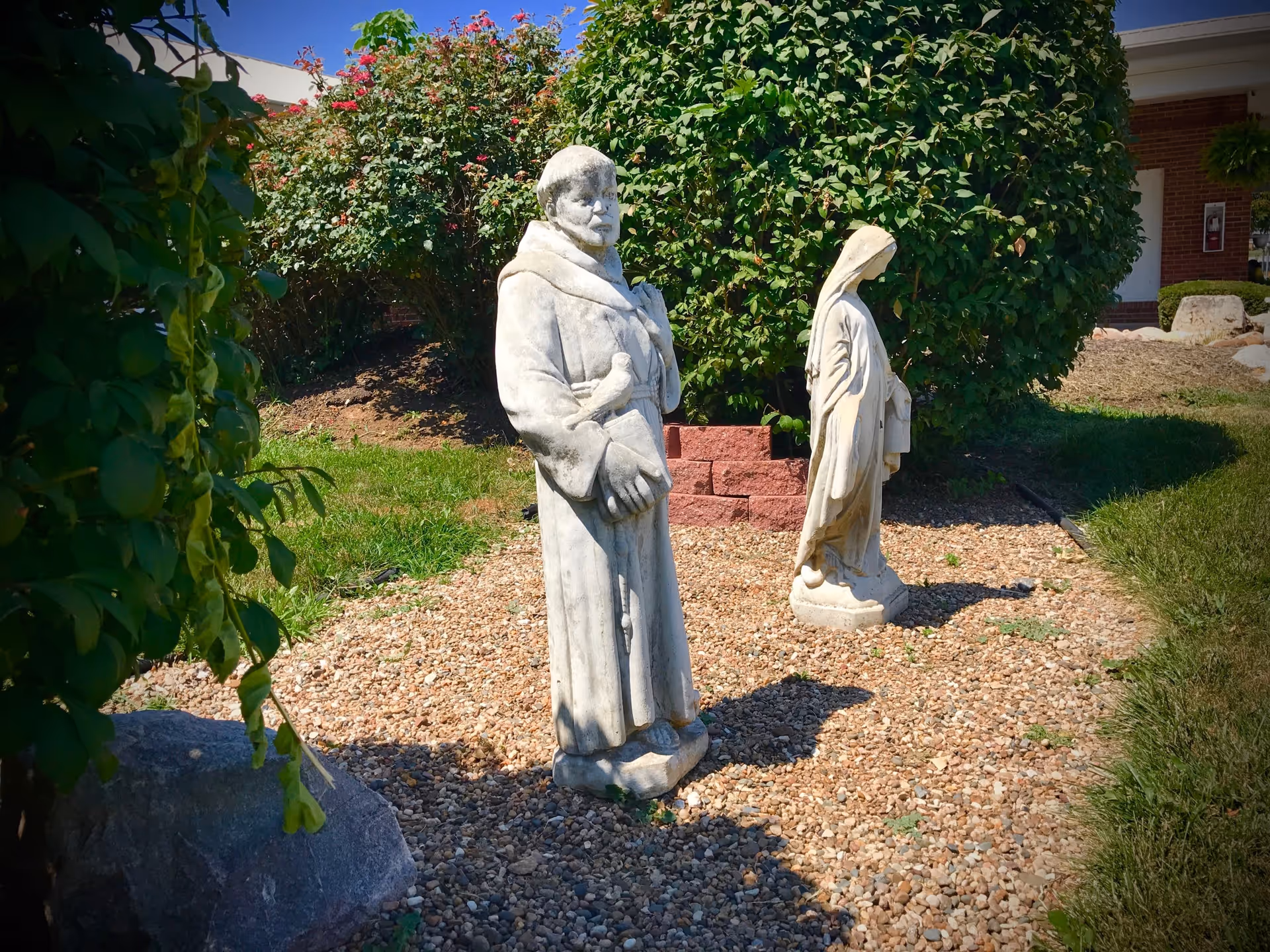 Two white stone statues in a garden area with gravel ground cover, surrounded by green bushes and grass. One statue depicts a man in a robe holding a bird and a book, while the other statue depicts a woman in a robe with a head covering. A building with a brick wall and a white door is visible in the background under a clear blue sky.