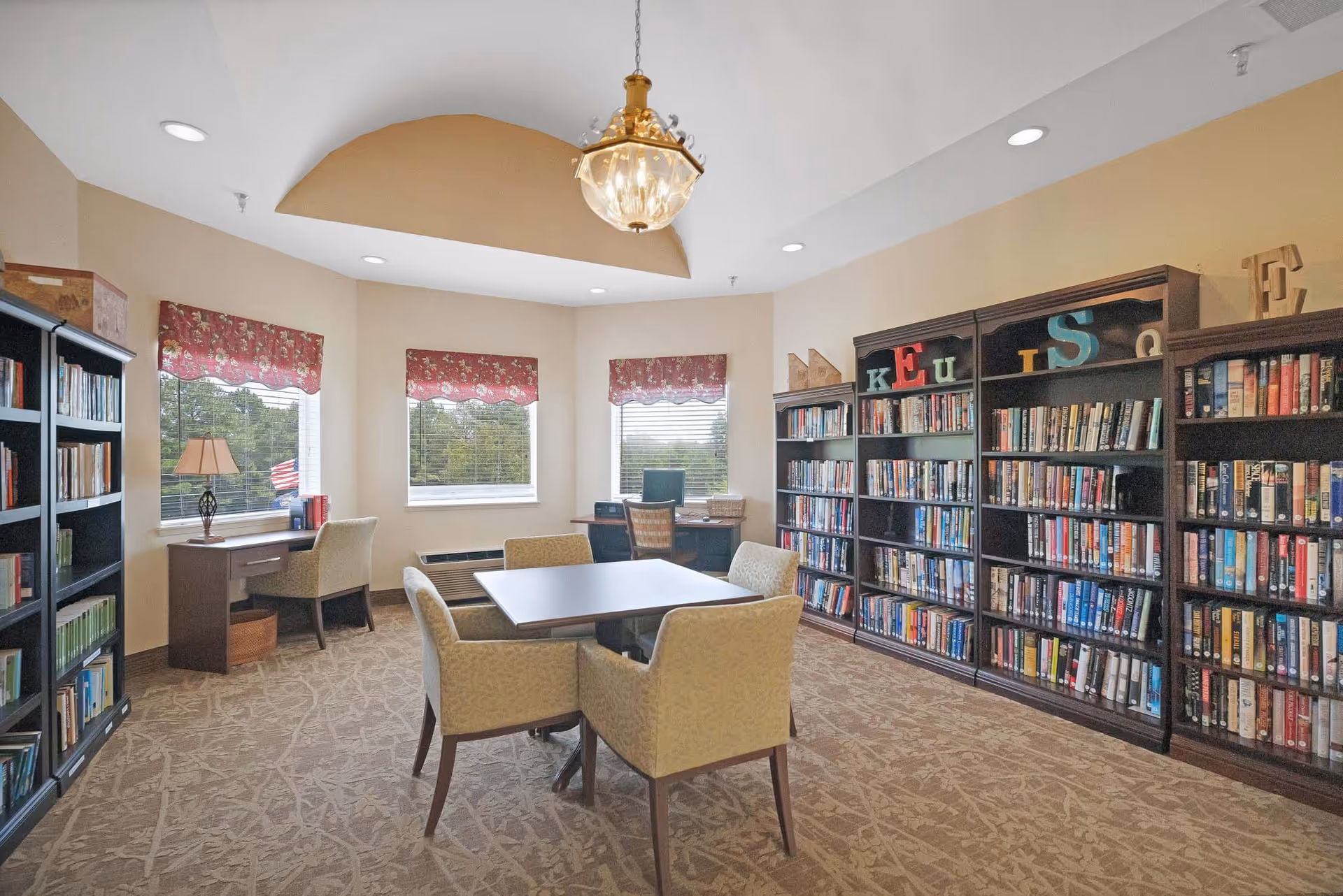 Well-lit interior reading room with bookshelves lining the walls, a central table with chairs, and desks beneath three windows.