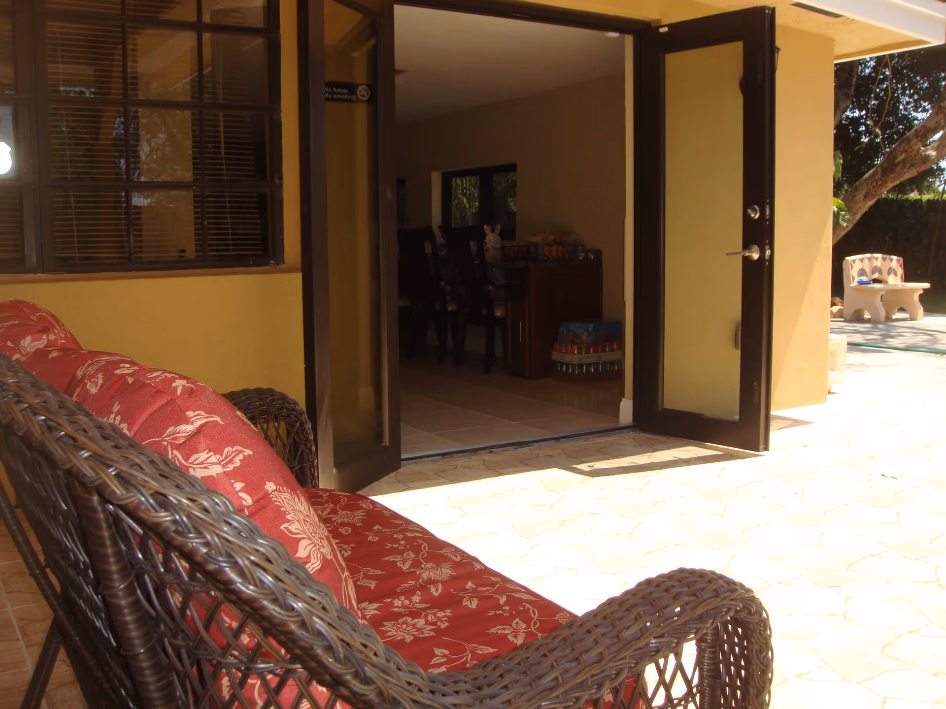 Outdoor patio area with a wicker sofa featuring red floral cushions. The patio has a stone floor and is adjacent to a building with open glass doors leading inside. Inside, a dining table with chairs and some stacked items are visible. There is a bench and a tree in the background outside.