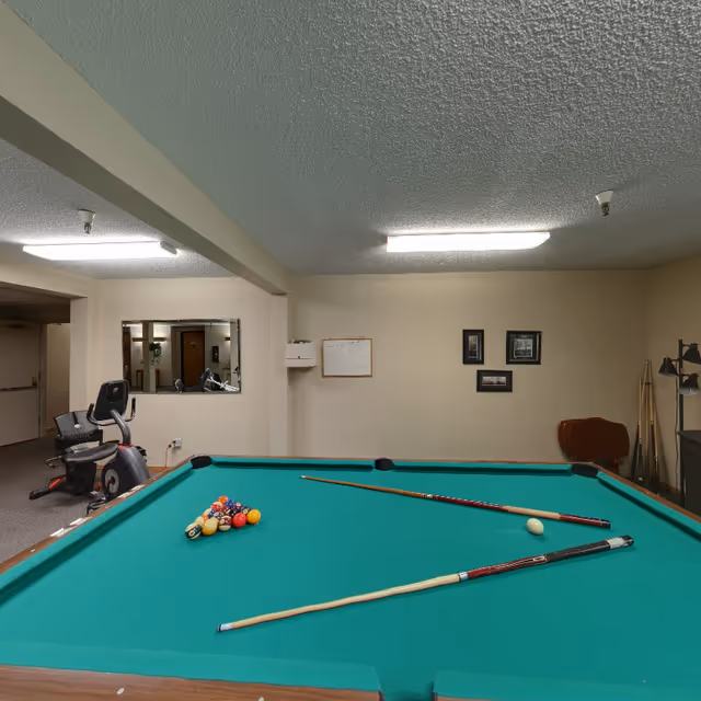 Interior room with a green pool table in the foreground, two pool cues and a rack of billiard balls on the table. In the background, there is an exercise bike, a mirror on the wall, a whiteboard, framed pictures, and a rack holding additional pool cues. The room has beige walls and a textured ceiling with fluorescent lighting.