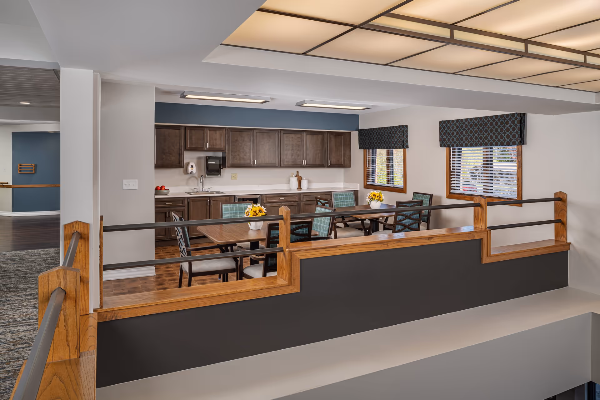 Open communal dining area with a kitchenette, table and chairs, and wooden railings overlooking a lower level.