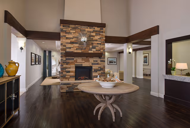 Interior view of a senior living facility featuring a central stone fireplace with a decorative sunburst mirror above it. In front of the fireplace is a round wooden table with a bowl of decorative items. The room has dark wood flooring, beige walls, and dark wood trim. To the left, there is a cabinet with decorative vases, and to the right, a reception or service counter with a lamp and a small plant. The space opens into other seating areas with framed artwork on the walls.
