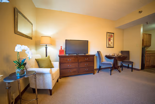 A cozy living area in a senior living facility featuring a beige armchair with a green pillow next to a small round table with a white orchid plant. A wooden dresser with a flat-screen TV on top is against the wall, accompanied by a floor lamp and a red decorative item. Two blue upholstered chairs are placed around a small round dining table set with dishes. The kitchen area is partially visible in the background.