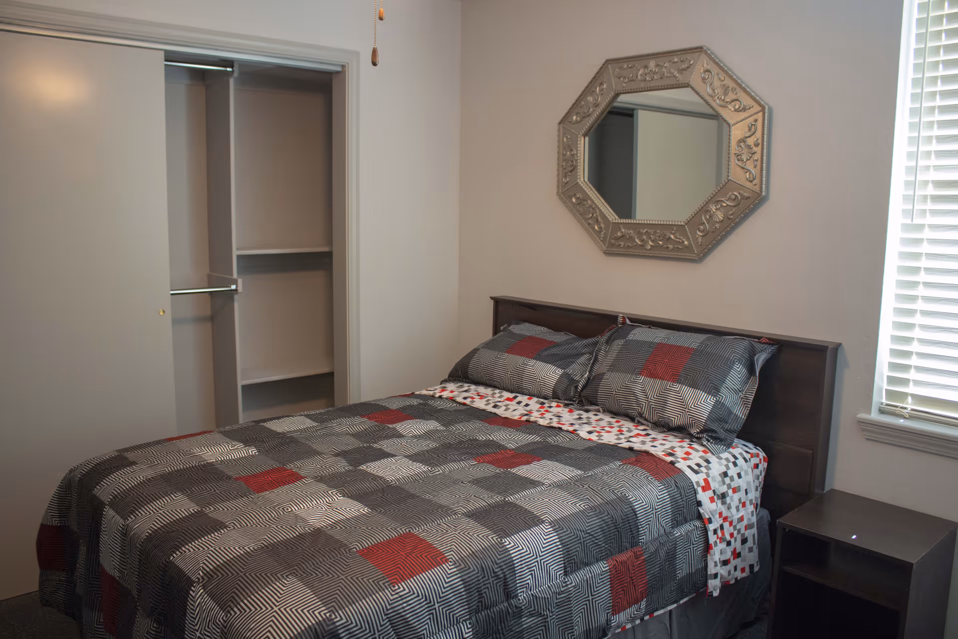 A bedroom with a bed covered in a patterned quilt featuring gray, black, and red squares. There are two matching pillows on the bed. Above the bed hangs an ornate octagonal mirror. To the right of the bed is a window with white blinds and a small dark wooden nightstand. On the left side of the room is an open closet with sliding doors.