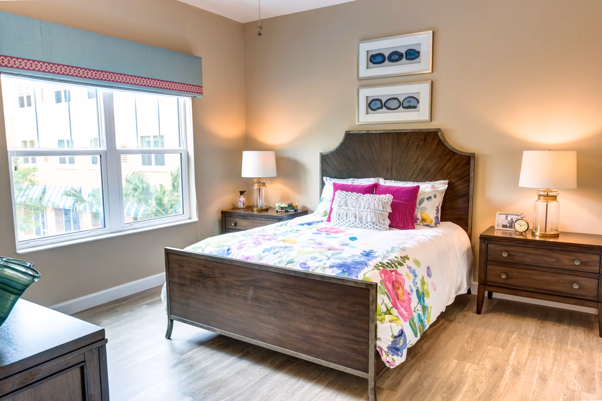 A bright and cozy bedroom with a wooden bed frame and floral bedding. The bed is adorned with decorative pillows in white and magenta. There are two wooden nightstands on either side of the bed, each with a lamp and small decorative items. A large window with a blue and pink valance lets in natural light, and two framed artworks hang above the headboard. The floor is light wood, and the walls are painted a soft beige.