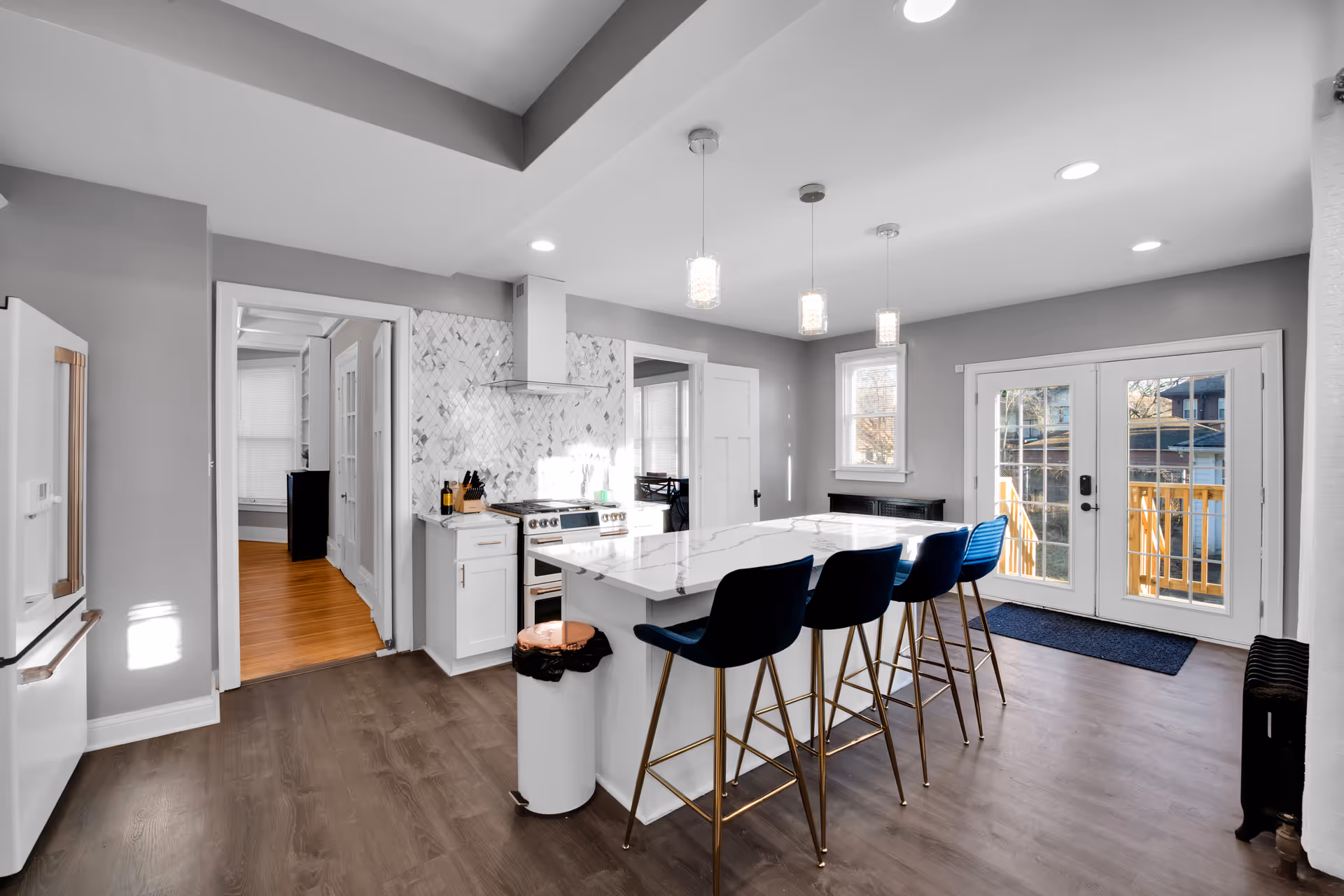 Bright modern kitchen with a marble island, four blue barstools, stainless appliances, pendant lights, and French doors to a deck.