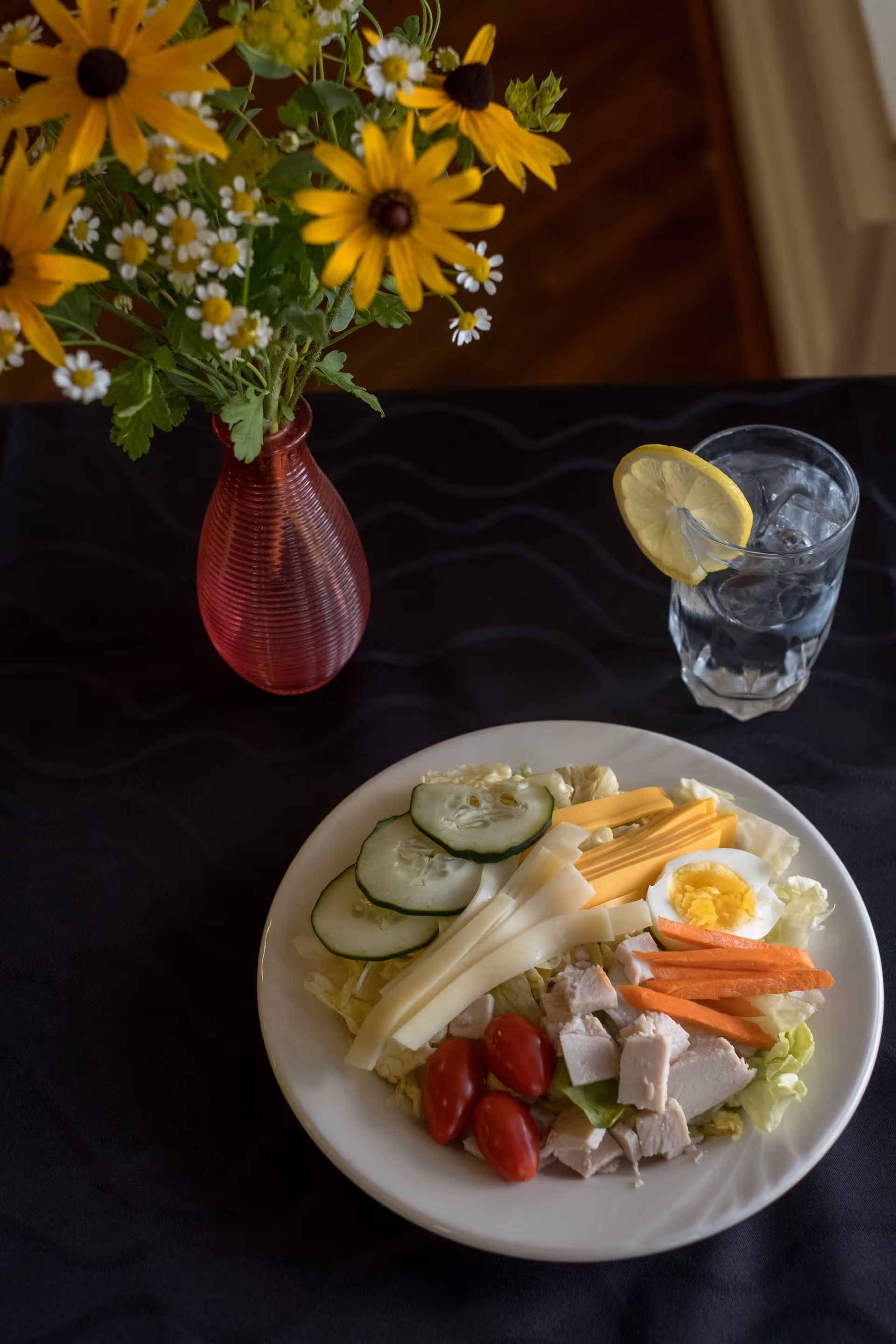 A plate of salad with sliced cucumbers, cheese sticks, cherry tomatoes, carrot sticks, chopped chicken, and half a boiled egg on a bed of lettuce. Next to the plate is a glass of water with a lemon slice on the rim. A red vase with yellow and white flowers is also on the black tablecloth.