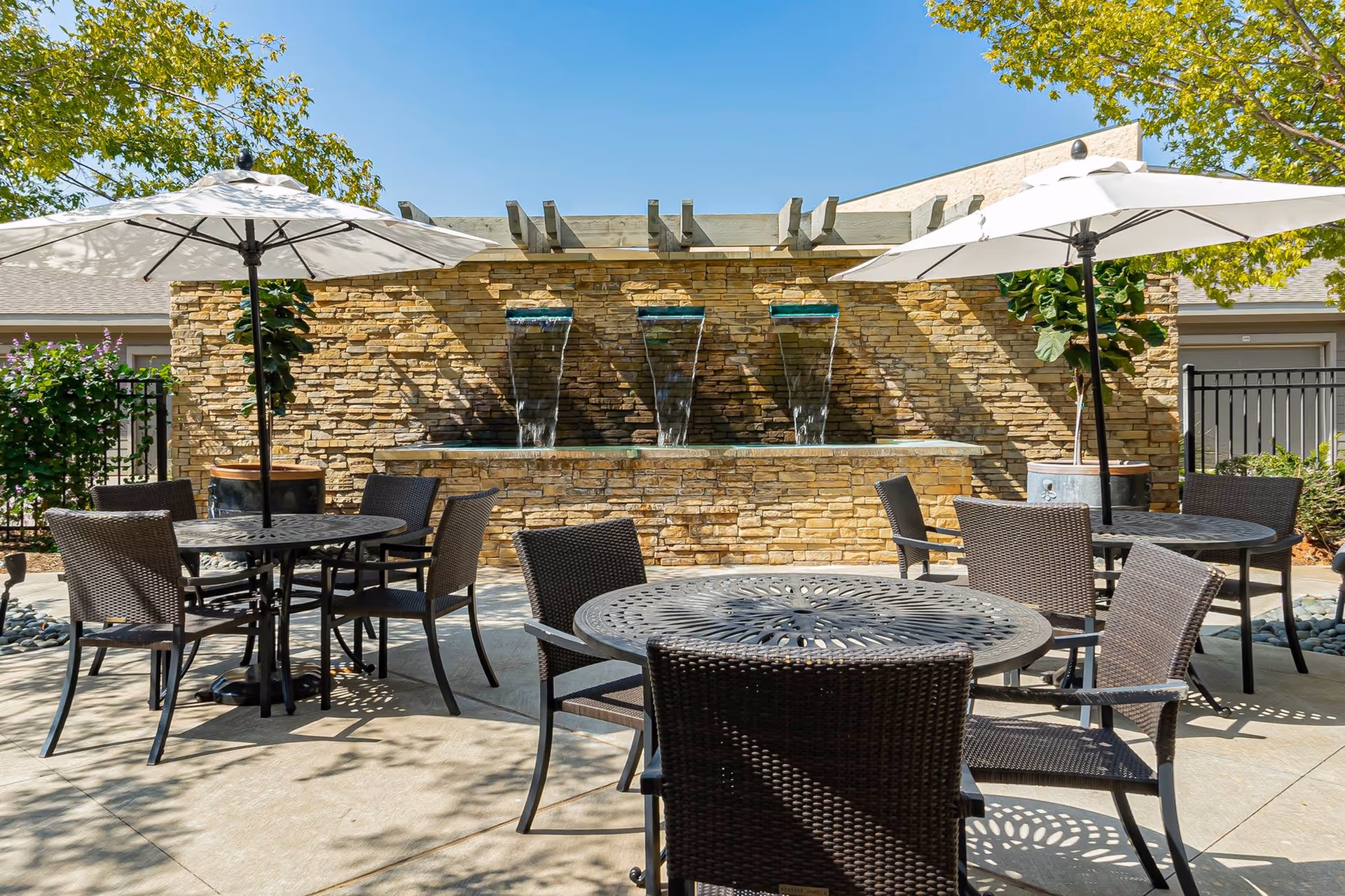 Outdoor patio area with round metal tables and wicker chairs under white umbrellas. A stone wall with three water fountains is in the background, surrounded by trees and plants under a clear blue sky.
