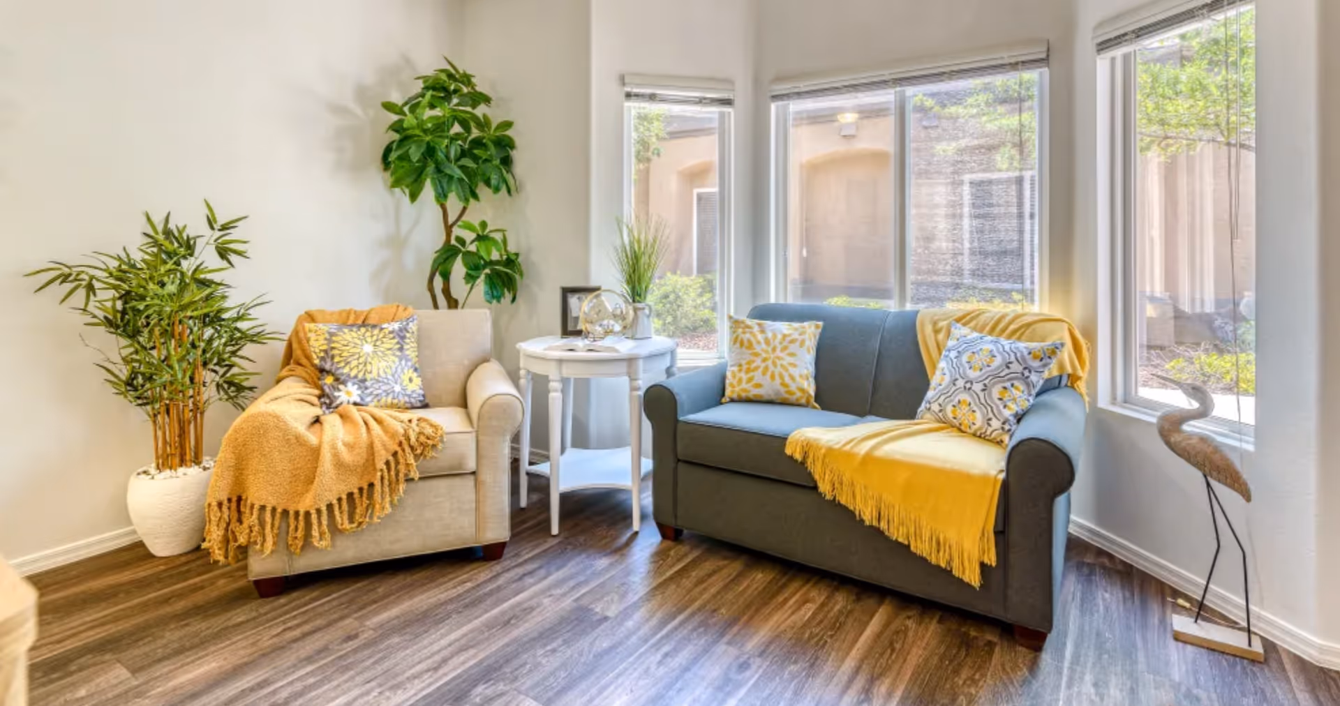 A cozy living room area with a beige armchair and a gray loveseat, both adorned with yellow and patterned throw pillows and yellow blankets. A small white round side table with decorative items is placed between the chairs. Large windows let in natural light, and there are two potted plants and a decorative bird sculpture near the windows.
