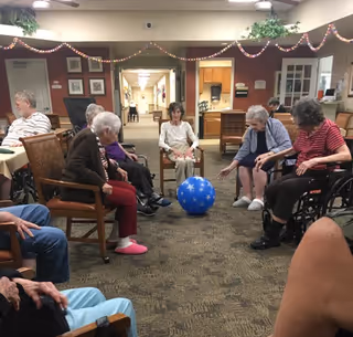 A group of elderly people sitting in a circle in a common area of an assisted living facility, engaging in a seated activity with a large blue ball with white stars. The room has carpeted floors, chairs arranged in a circle, and decorations hanging from the ceiling. There is a hallway and a kitchenette visible in the background.