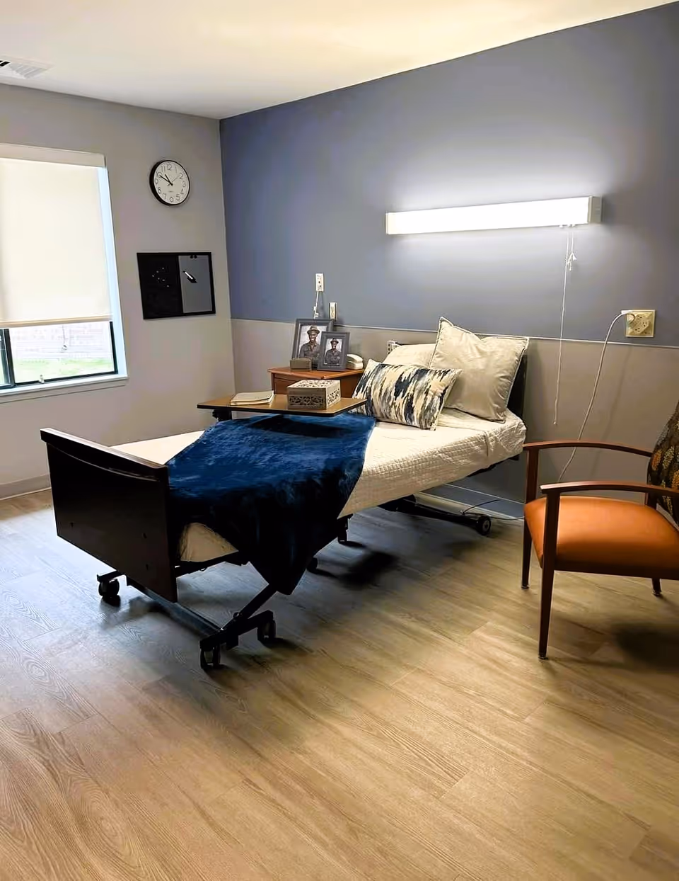 A tidy nursing facility bedroom with a single adjustable bed, bedside table with framed photos, an armchair and a window.