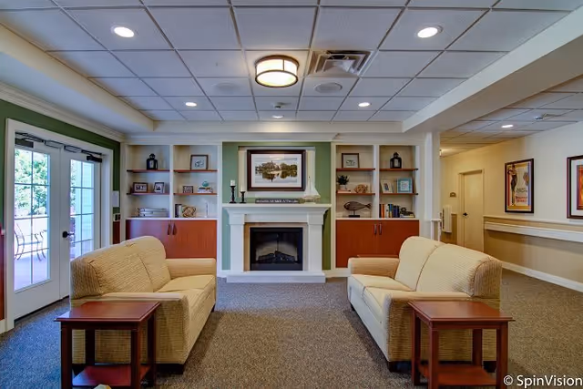 A cozy lounge with two beige sofas facing a fireplace flanked by built-in shelves, French doors to the left and ceiling lights above.