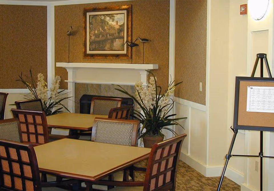 Interior view of a common area with multiple tables and chairs arranged for seating. There is a white fireplace with a framed painting above it, decorative bird sculptures on the mantel, and potted plants with white flowers on either side. A bulletin board on an easel stands to the right against a beige and white paneled wall.