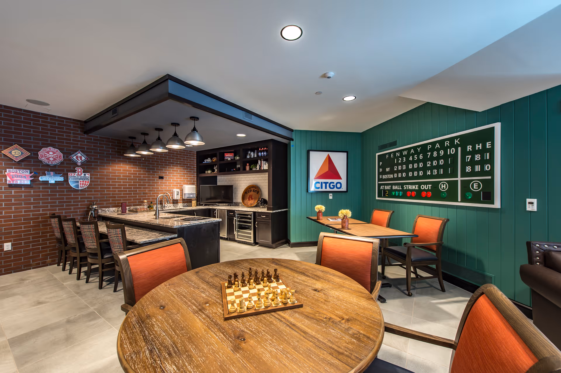 A cozy interior room featuring a kitchen area with a granite countertop and bar stools, a round wooden table with a chessboard set up, and a smaller table with orange chairs. The walls are decorated with Boston Red Sox memorabilia, including a Fenway Park scoreboard and a Citgo sign. The room has a warm and inviting atmosphere with brick and green paneled walls.