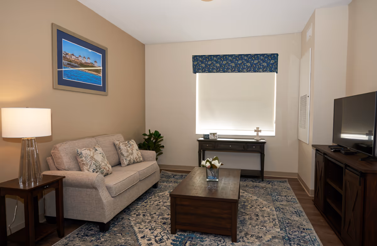 Neatly furnished living room with a sofa, coffee table, TV console, lamp, and a window with a valance.