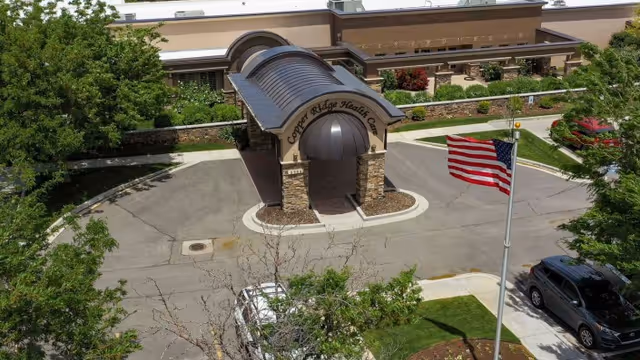 Aerial view of the entrance to Copper Ridge Health Care facility featuring a covered drop-off area with stone pillars and a curved roof. The building is surrounded by greenery, a parking lot with cars, and an American flag on a flagpole.