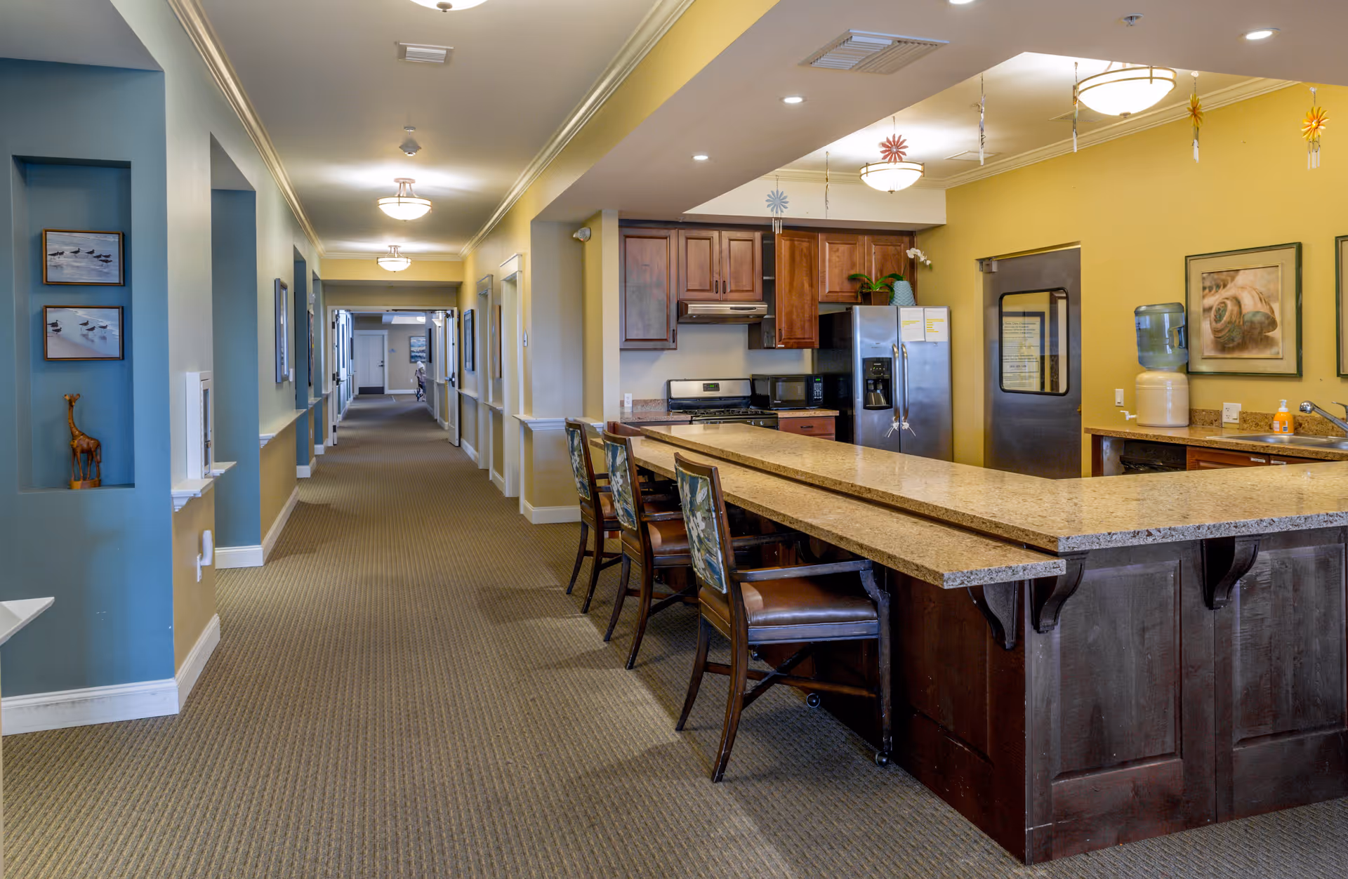 Interior view of a memory care facility hallway with a kitchen area on the right. The kitchen features a granite countertop with several chairs, wooden cabinets, a refrigerator, stove, microwave, and a water dispenser. The hallway is carpeted and decorated with framed pictures and small sculptures.