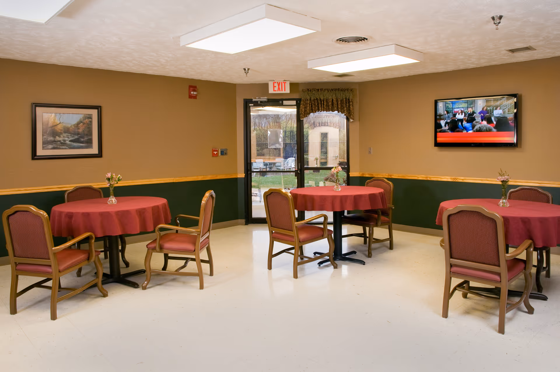 A dining area with three round tables covered with red tablecloths, each surrounded by four wooden chairs with red cushions. Small flower vases are placed on each table. The room has beige and green walls, a framed landscape painting, a wall-mounted TV showing a group of people, and a glass door leading outside.