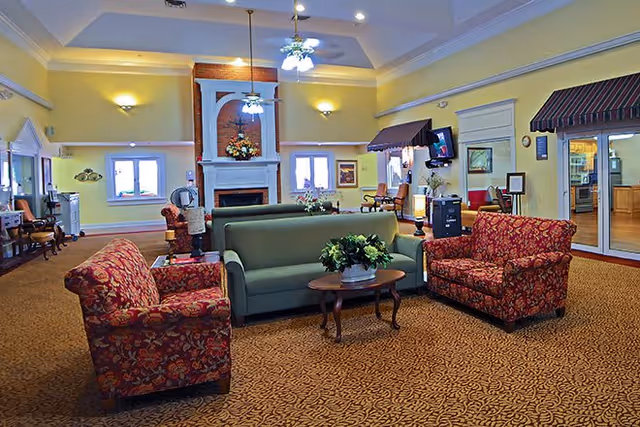 A spacious living room area with yellow walls and a high ceiling featuring crown molding. The room has a green sofa and two red floral armchairs arranged around a wooden coffee table with a flower arrangement. There is a fireplace with a decorative mantel and floral arrangement above it. The room is well-lit with ceiling fans and wall sconces. Large windows and glass doors allow natural light to enter, and there are various pieces of furniture and decor throughout the space.