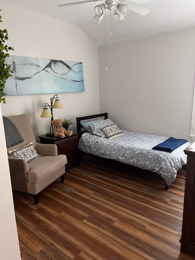 A small, neatly arranged bedroom with a single bed covered in a patterned gray and white bedspread, a beige armchair with a decorative pillow, a wooden nightstand with a lamp and a teddy bear, a piece of abstract wall art above the nightstand, and a ceiling fan with lights. The room has wooden flooring and white walls.