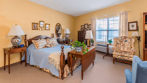 A cozy bedroom with yellow walls featuring a wooden bed with a blue bedspread and multiple pillows. There are two bedside tables with decorative lamps, a wooden table with a potted plant at the foot of the bed, a floral upholstered armchair near a window with light curtains, and framed pictures on the walls.