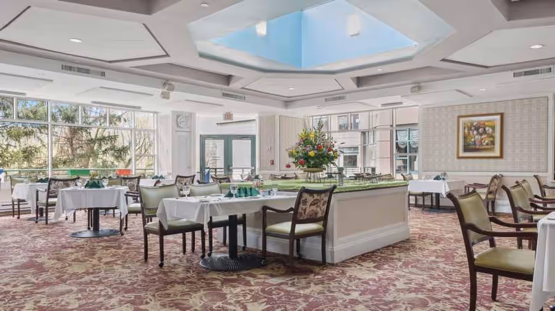 A spacious dining room with multiple tables covered in white tablecloths, set with glassware and napkins. The room features large windows allowing natural light to fill the space, a floral centerpiece on a central table, patterned carpet, and a skylight in the ceiling. There is a framed painting on the wall and an exit door visible in the background.