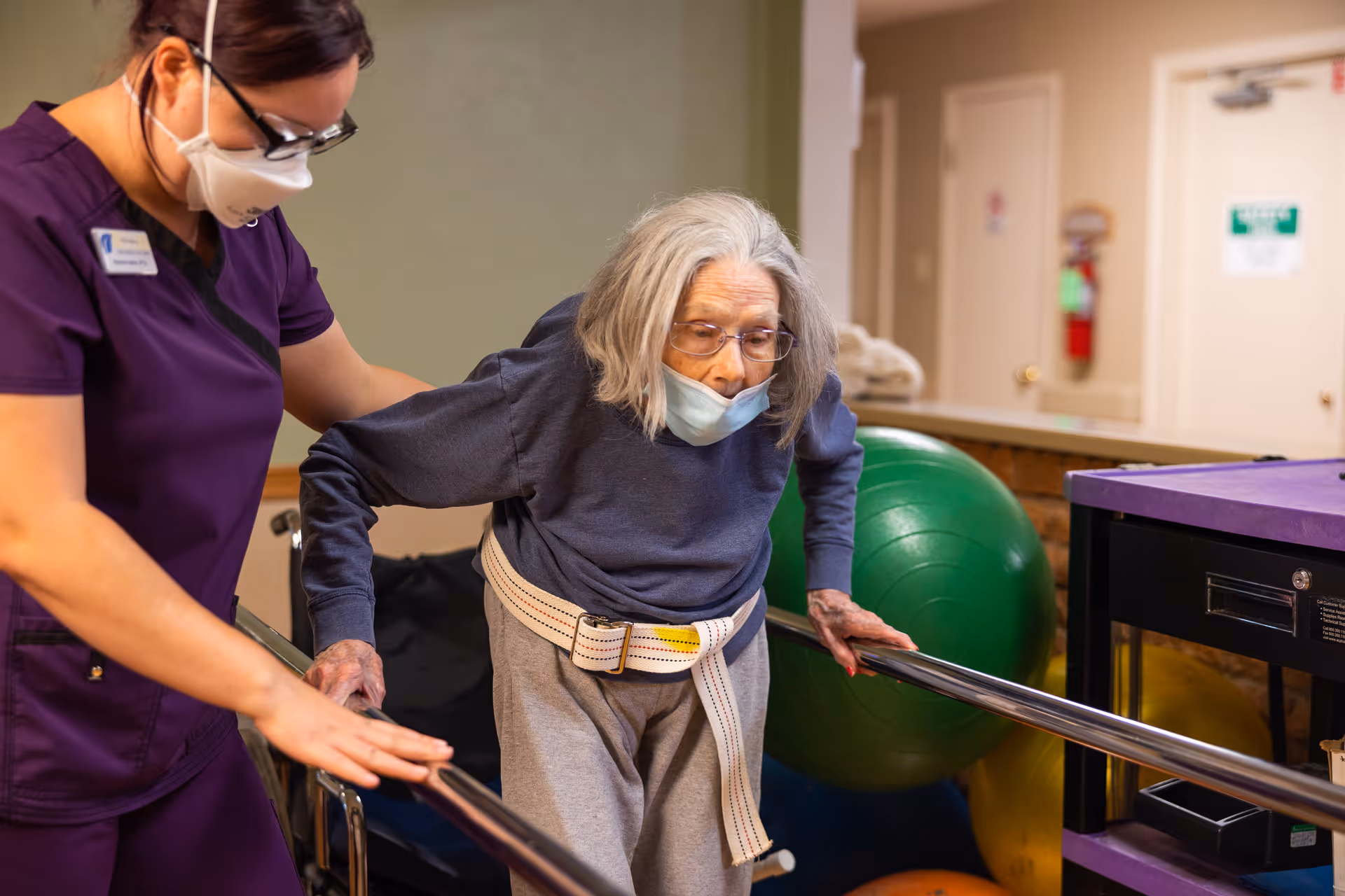 A healthcare worker wearing a purple uniform and face mask assists an elderly woman with gray hair and glasses, who is also wearing a face mask and a gait belt, as she practices walking between parallel bars in a rehabilitation setting with exercise balls in the background.