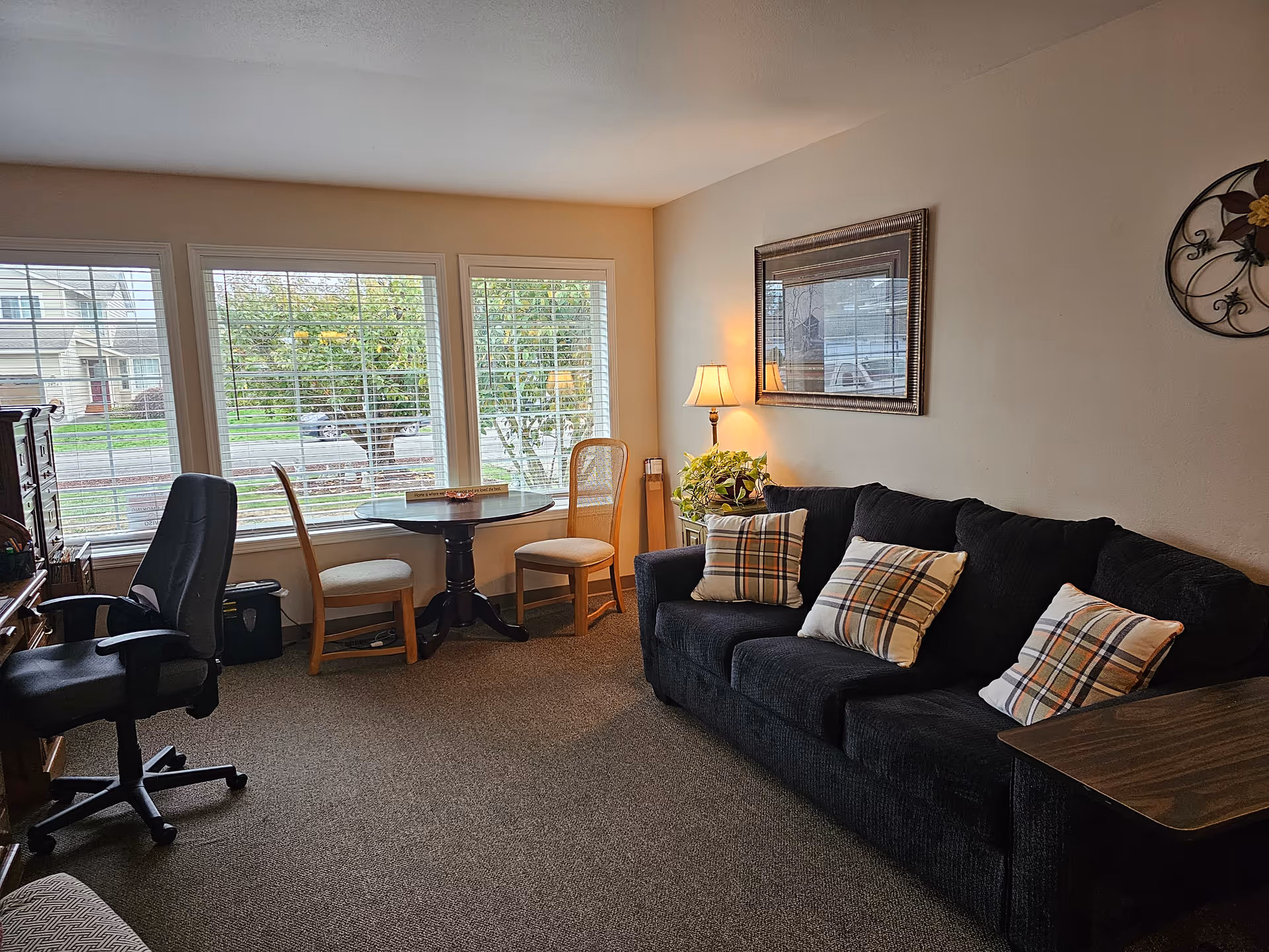 Sunlit living room with a dark sofa with plaid pillows, a round table by three large windows and an office chair.