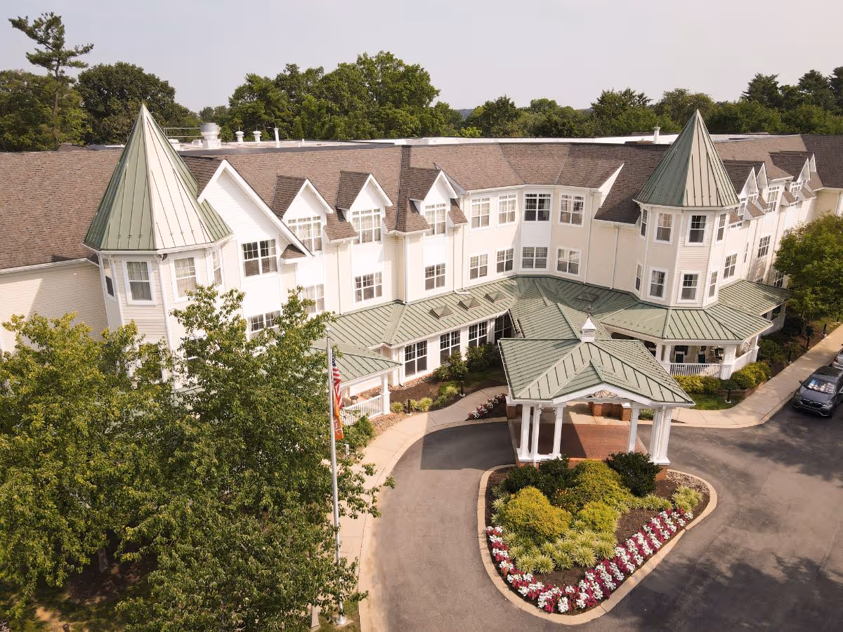 Aerial view of the exterior front entrance of a senior living facility named Sunrise of Lafayette Hill, featuring a large white building with green peaked roofs, multiple windows, and a covered driveway entrance surrounded by landscaped bushes and flowers.