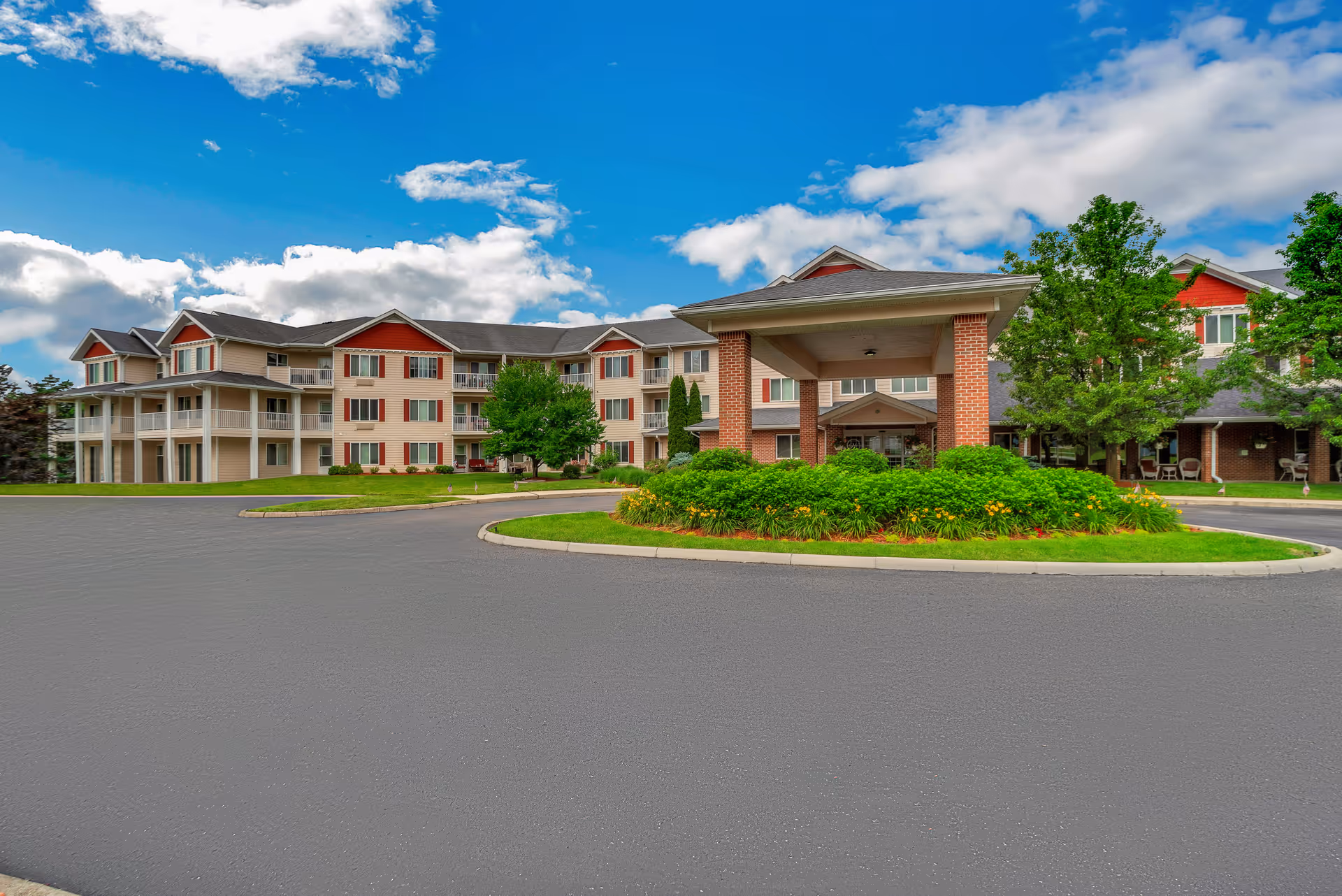 Front exterior of a multi-story senior living facility with a covered entrance, landscaped roundabout, balconies, and trees under a blue sky.