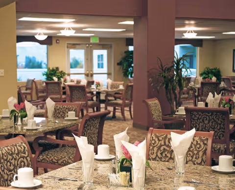 A spacious dining room with multiple set tables and chairs, napkins and tableware, and decorative plants and columns.
