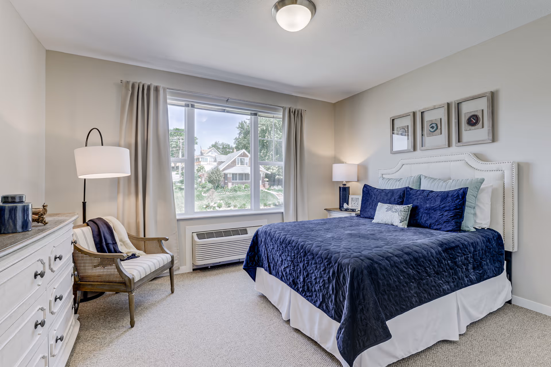 A bright and cozy bedroom with a large window showing a view of houses and greenery outside. The room features a bed with a navy blue quilt and multiple pillows, a white headboard, a wooden chair with a cushion and throw blanket, a white dresser with decorative items, and two lamps providing warm lighting.