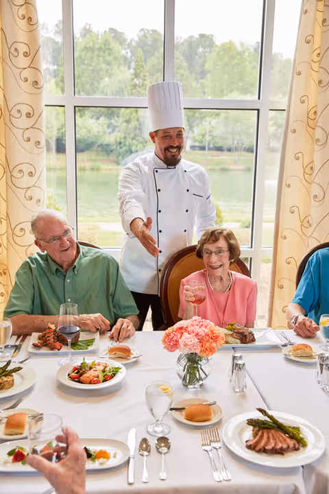 A chef stands behind smiling elderly diners at a table set with plates, glasses, and a vase of flowers in front of a large window.