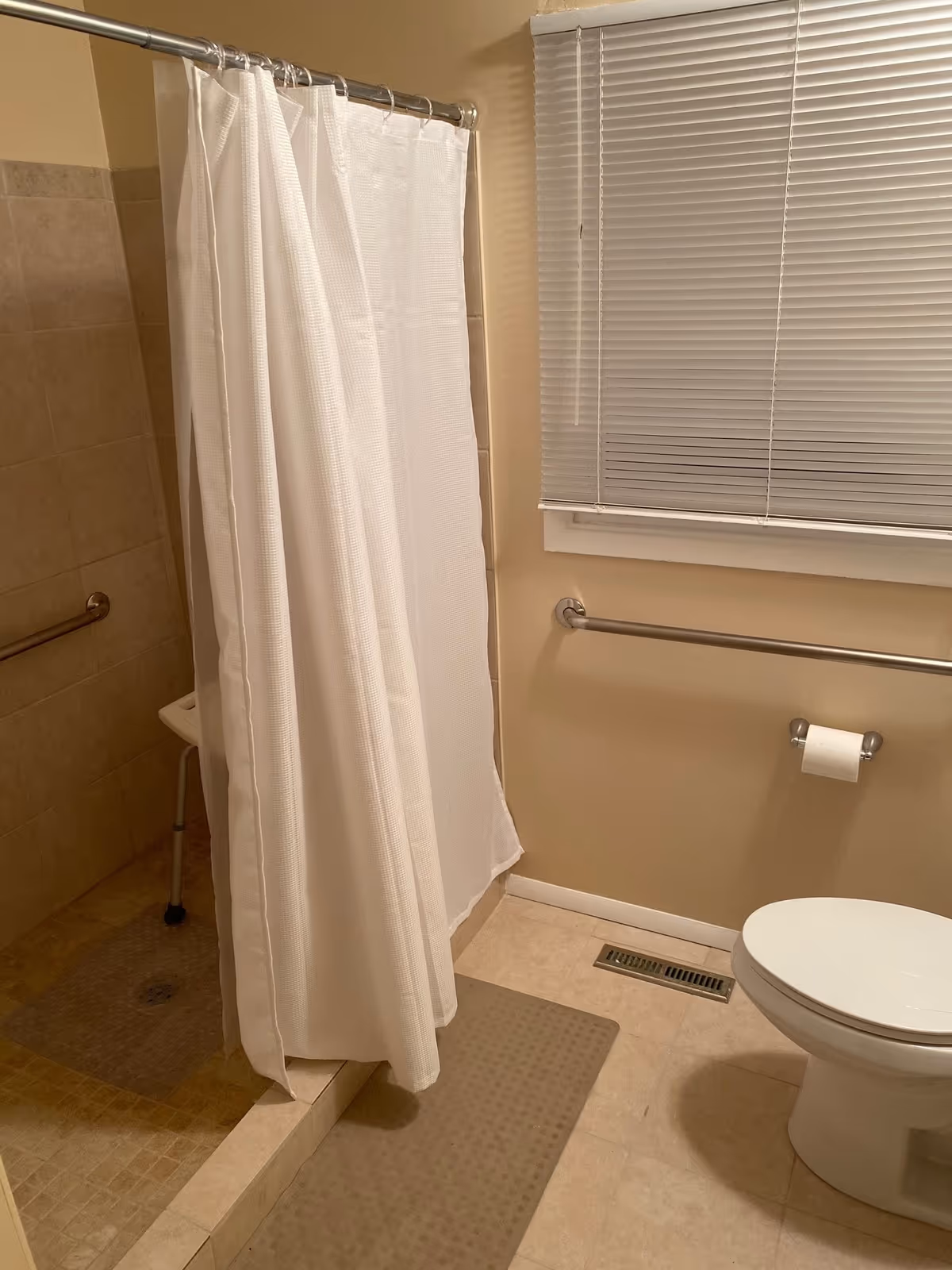 A bathroom with a walk-in tiled shower featuring a white shower curtain and a shower chair inside. There is a toilet with a toilet paper holder mounted on the wall next to it, a grab bar on the wall, a window with closed blinds, and a floor vent visible on the tiled floor.