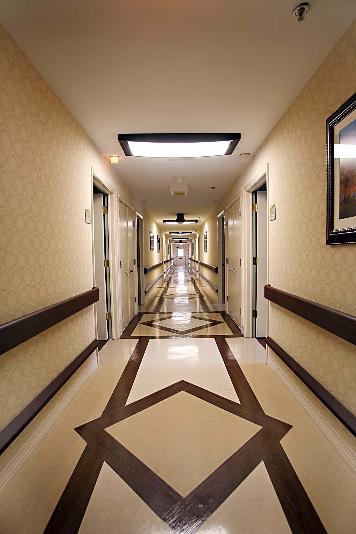 Long hallway in a senior living facility with patterned beige walls, wooden handrails on both sides, multiple closed doors, framed artwork on the walls, and geometric patterned flooring. Ceiling lights illuminate the corridor.
