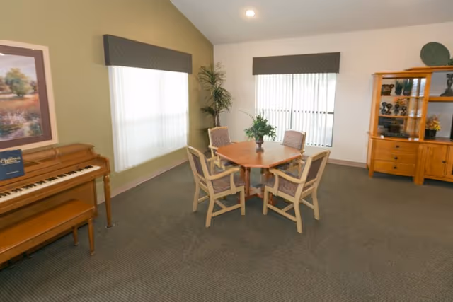 Communal sitting room with a round wooden table and four chairs, a piano on the left, and a wooden cabinet on the right.