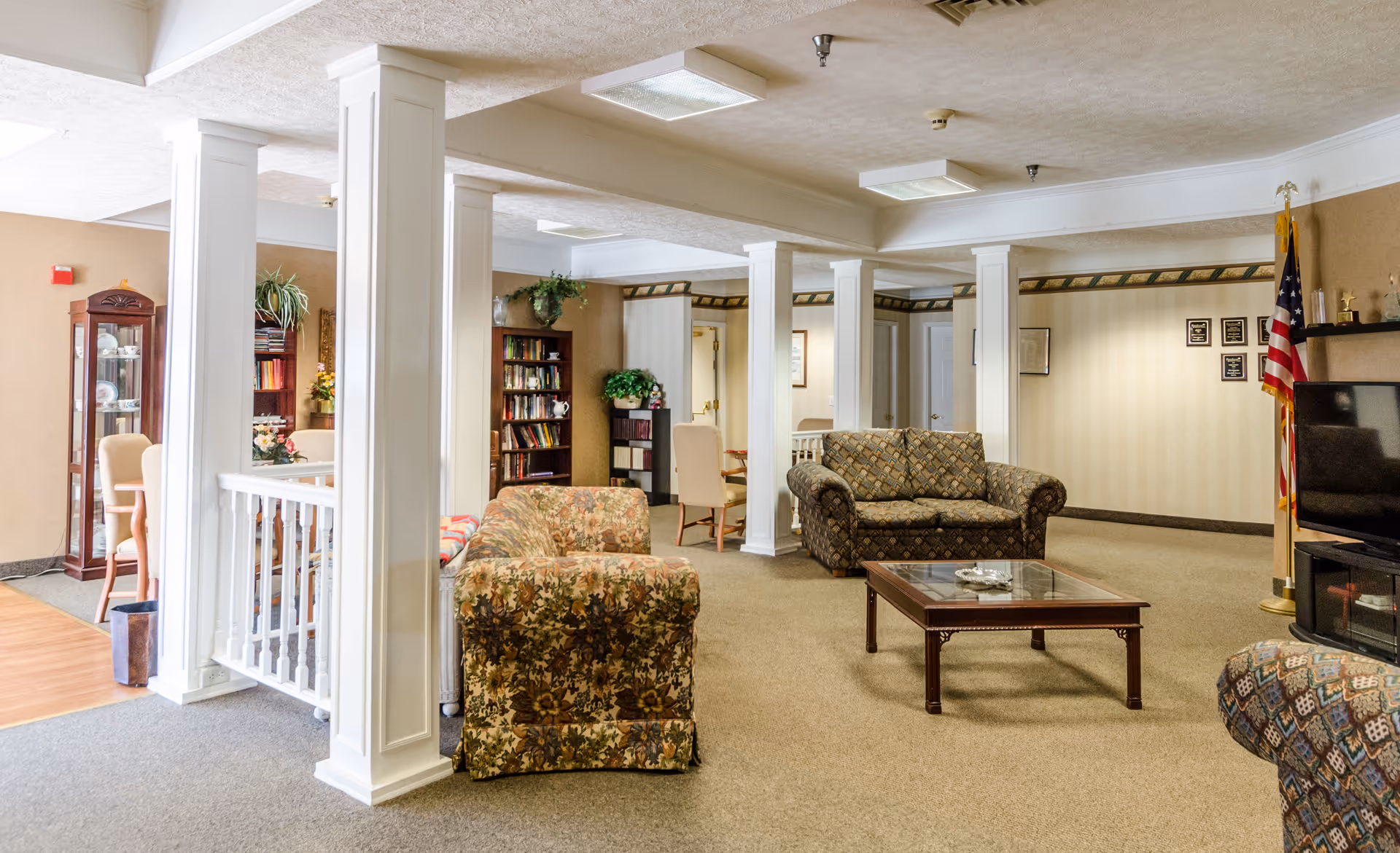 A spacious communal living room with upholstered sofas and armchairs, a coffee table, bookshelves, decorative columns, and an American flag.