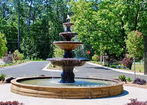 A large multi-tiered stone fountain in a paved circular drive surrounded by trees and landscaping.