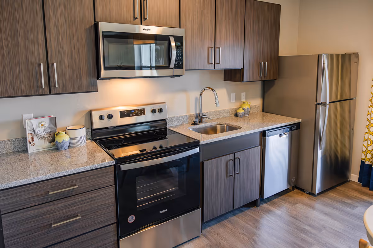 Modern kitchen with stainless steel appliances including a microwave, stove, dishwasher, and refrigerator. The kitchen features wood grain cabinets, a granite countertop with a sink and faucet, and decorative items such as a cookbook, bowls, and vases.