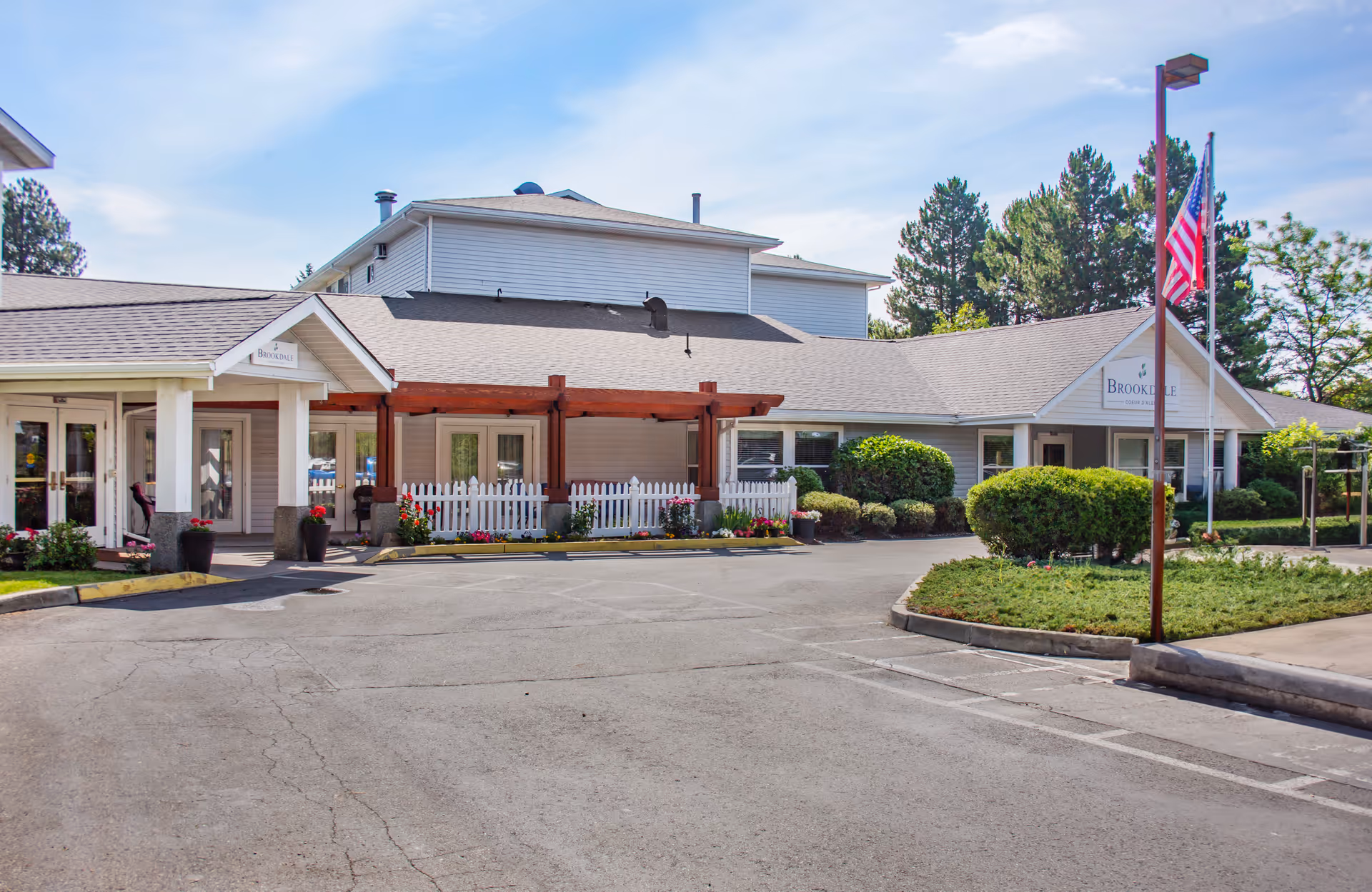 Front entrance of the Brookdale Coeur d'Alene building with a covered drop-off, flagpole, white picket fence, and landscaped shrubs.