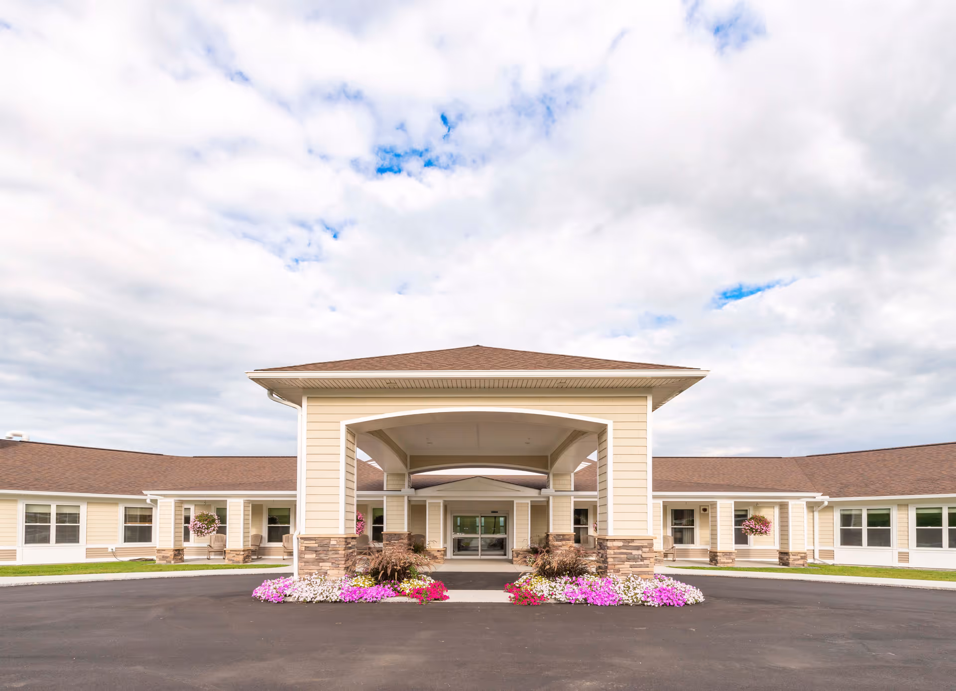 Front exterior view of a single-story senior living facility building with beige siding, stone accents, a covered entrance, and colorful flower beds in front under a partly cloudy sky.