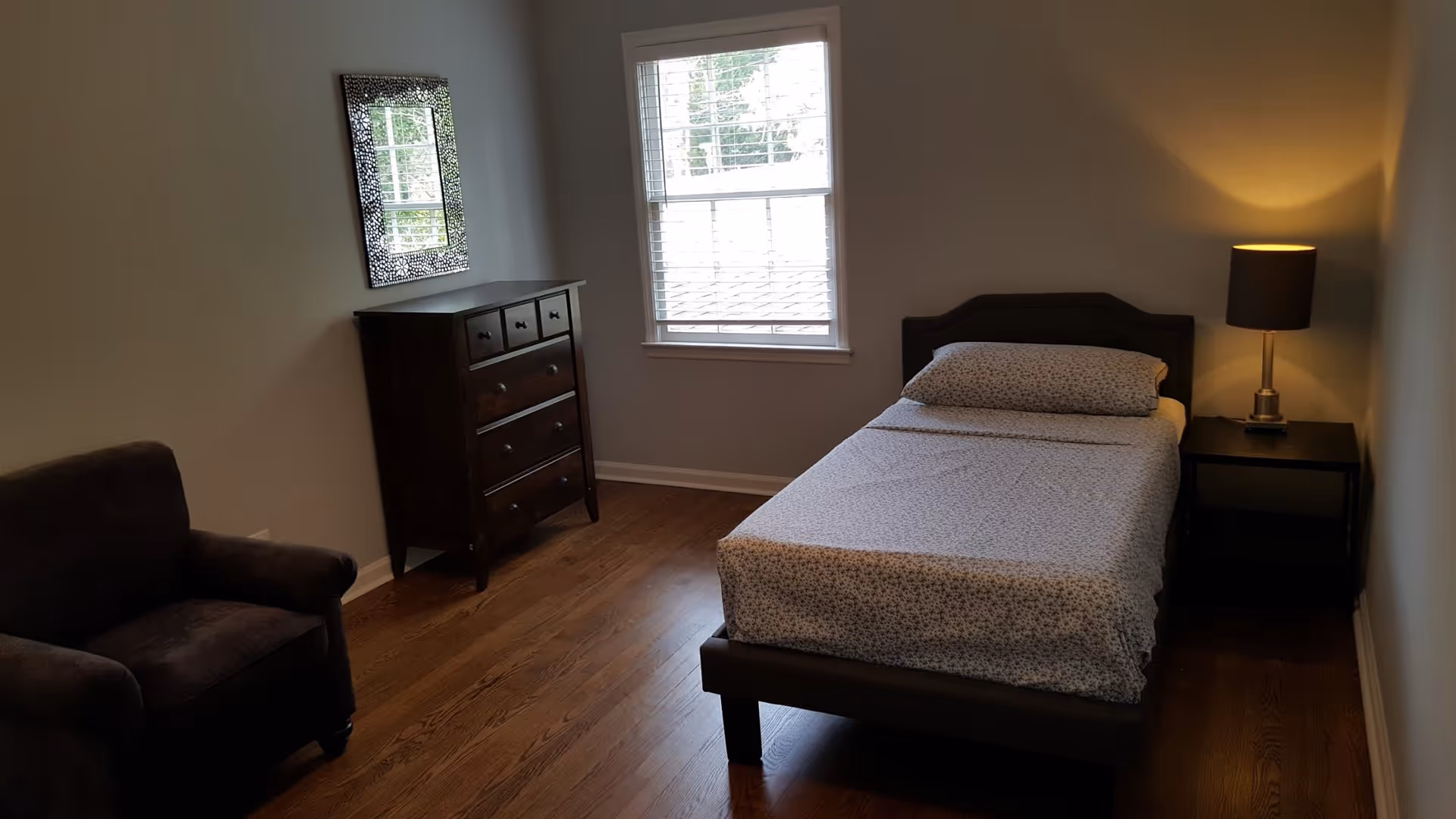 A simple bedroom with a single bed covered in patterned sheets, a dark wooden dresser with a decorative mirror above it, a dark armchair, a window with white blinds, and a nightstand with a lit lamp.